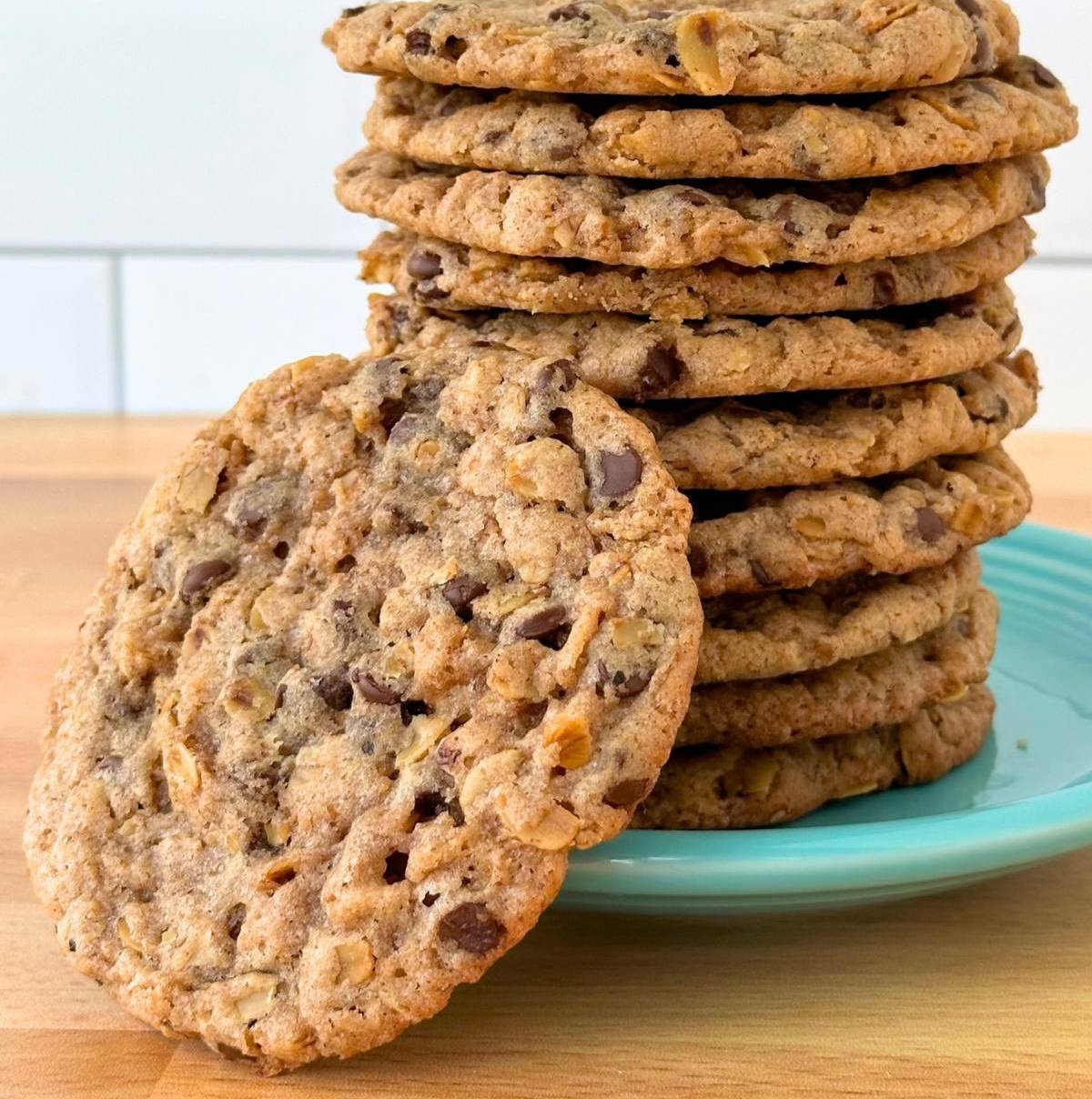 A stack of homemade cookies with chocolate chips and nuts is neatly arranged on a turquoise plate. One cookie is leaning against the stack, showcasing its texture with visible oats and chocolate chips. The background is a light-colored countertop with whi
