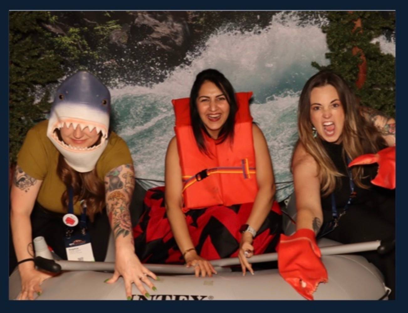 Three women in life raft. One woman is wearing a shark helmet.