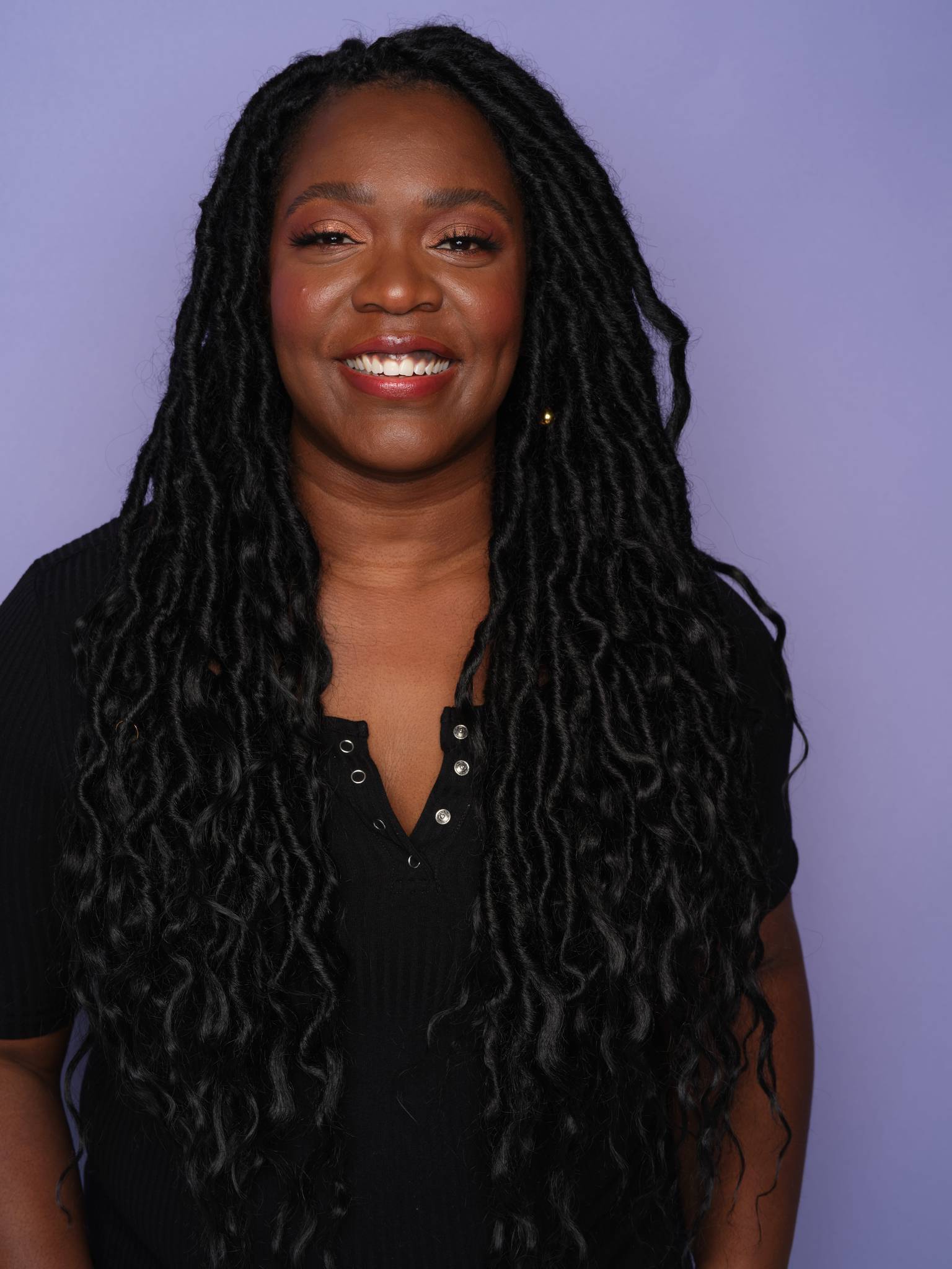 Alexandra Kane stands, with a warm and inviting smile. She has long locs and a black tshirt on. 