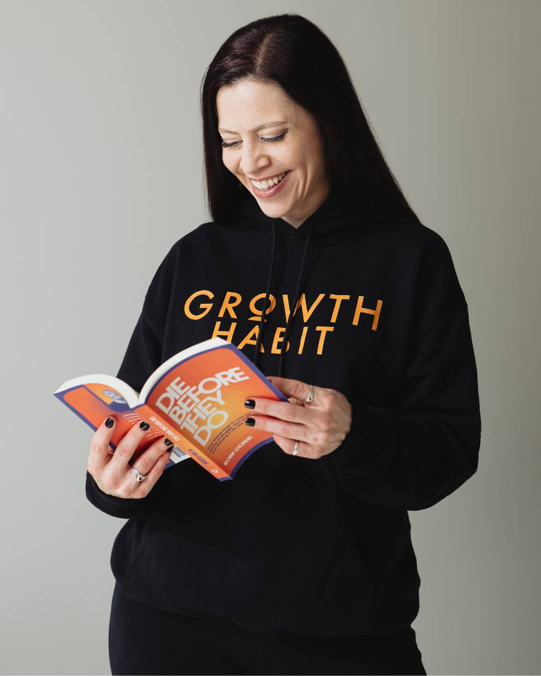 Moriah wearing a black Growth Habit hoodie and holding a copy of Die Before They Do