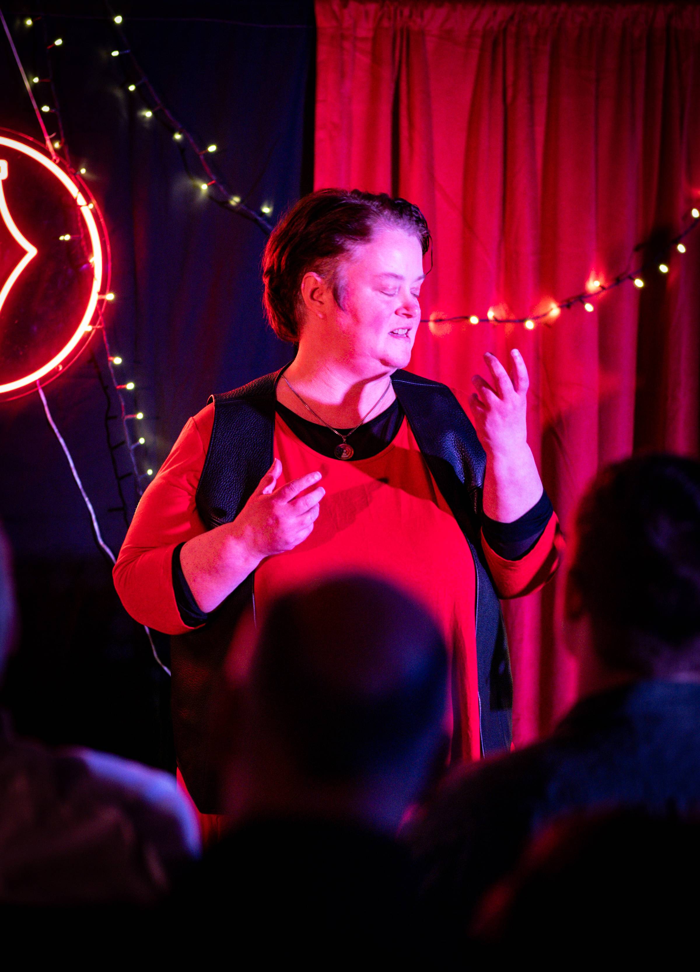 Joanna Evervenn gesturing to the side and looking at their hands while wearing red and black against a red and black background. Image by Fleassy Malay.