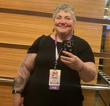 Selfie of Laurie taken at the Seattle 2024 Fat Con. Laurie is a white, mid-fat woman with short grey and brown hair wearing dangling earrings and a black short sleeve dress.