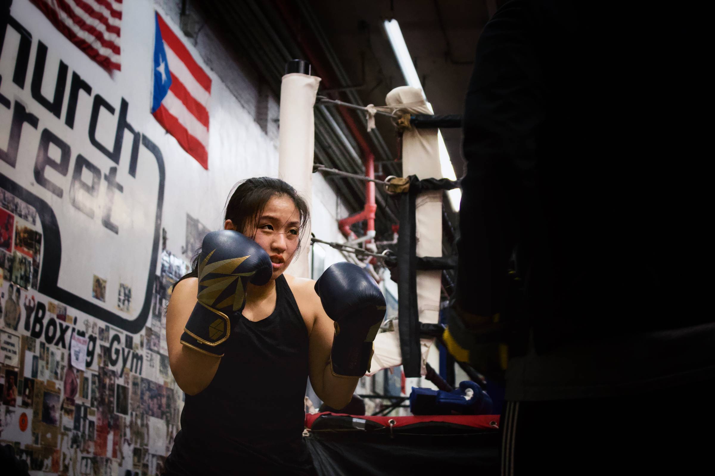 Asian woman wearing all black in a boxing gym with gloved hands held near her face.