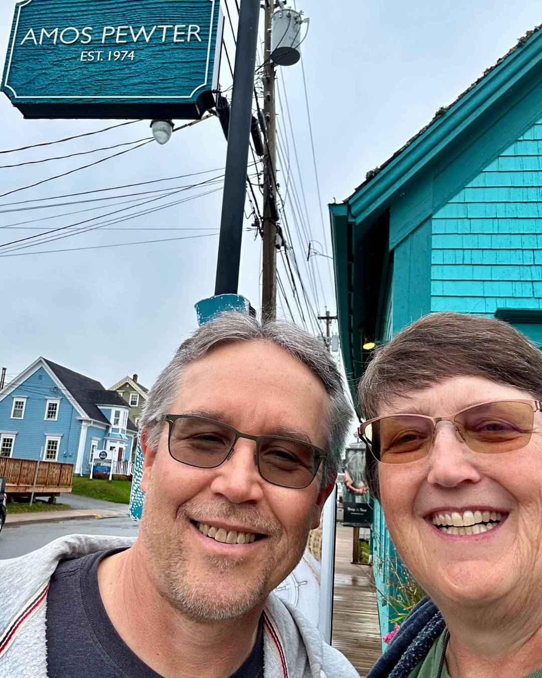 A man and woman smiling at the camera with a teal building behind them and a sign with "AMOS PEWTER."