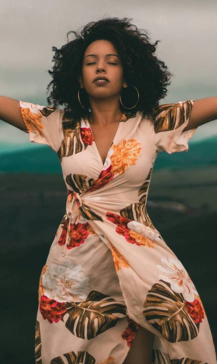 Black woman with arms open wide and black curls blowing in wind wearing a floral wrap dress