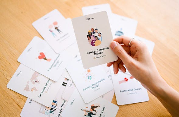 A person holding a card that says equity-centered design. In the background with a slight blur, we see a pile of cards that say restorative design, socially responsible design, co-creation and trauma-informed design