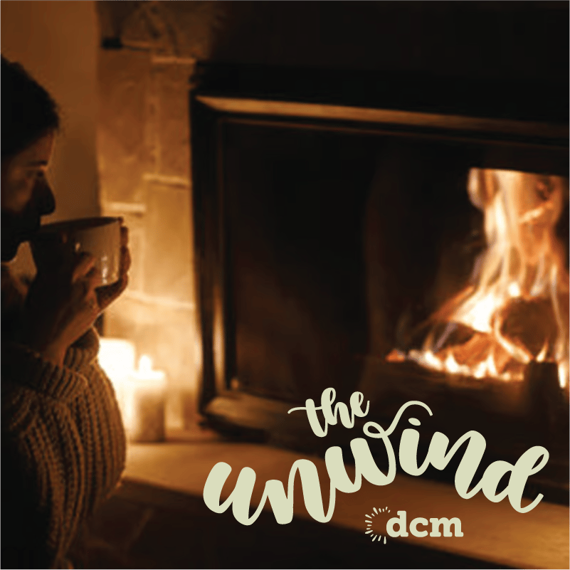 Person in a cozy sweater sipping a mug beside a lit fireplace and candles; overlaid text reads “the unwind” and “dcm.”