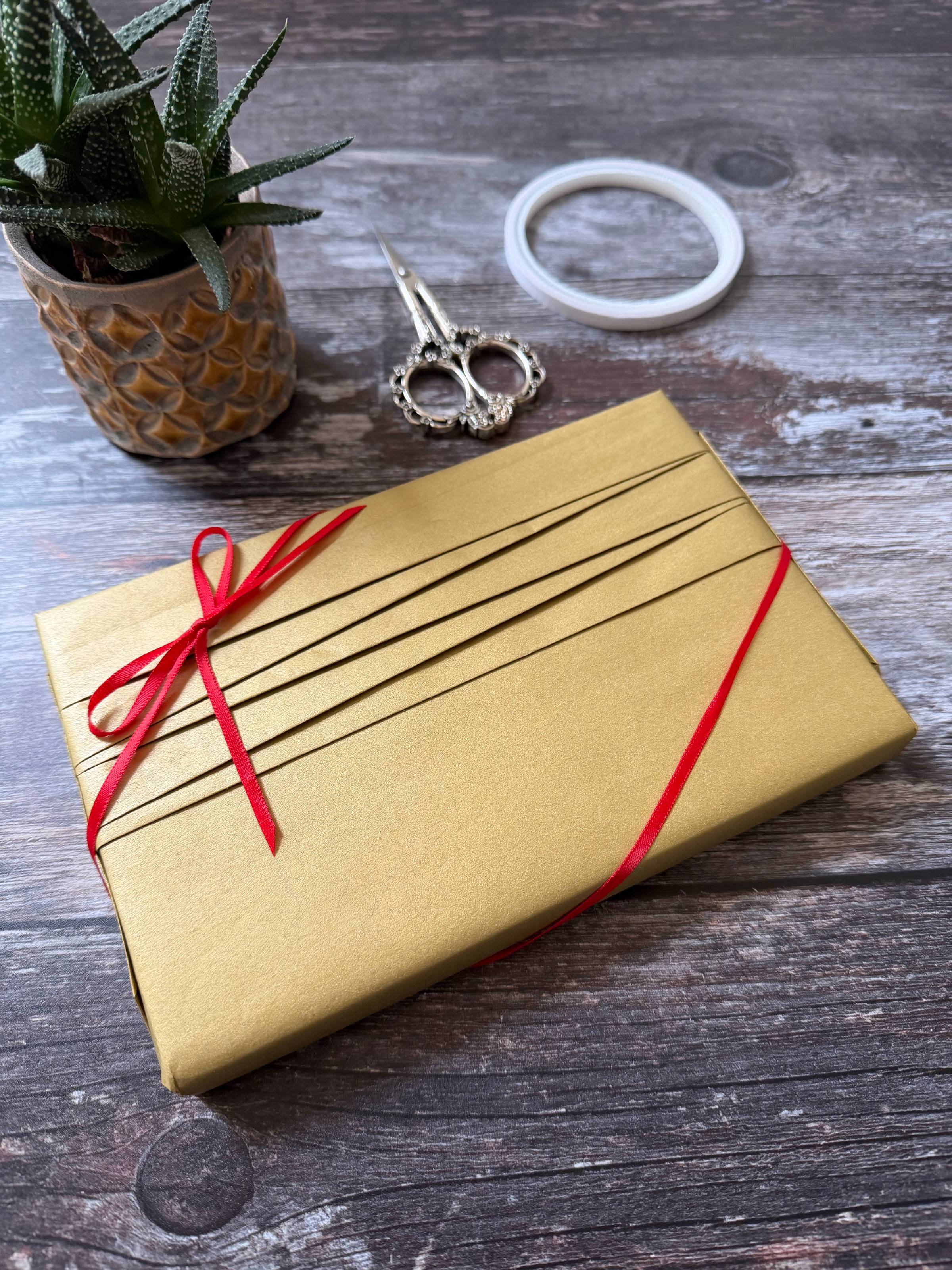 Gold wrapping paper with a folded pleating effect on the front and a narrow red ribbon tied in a bow at the corner. 