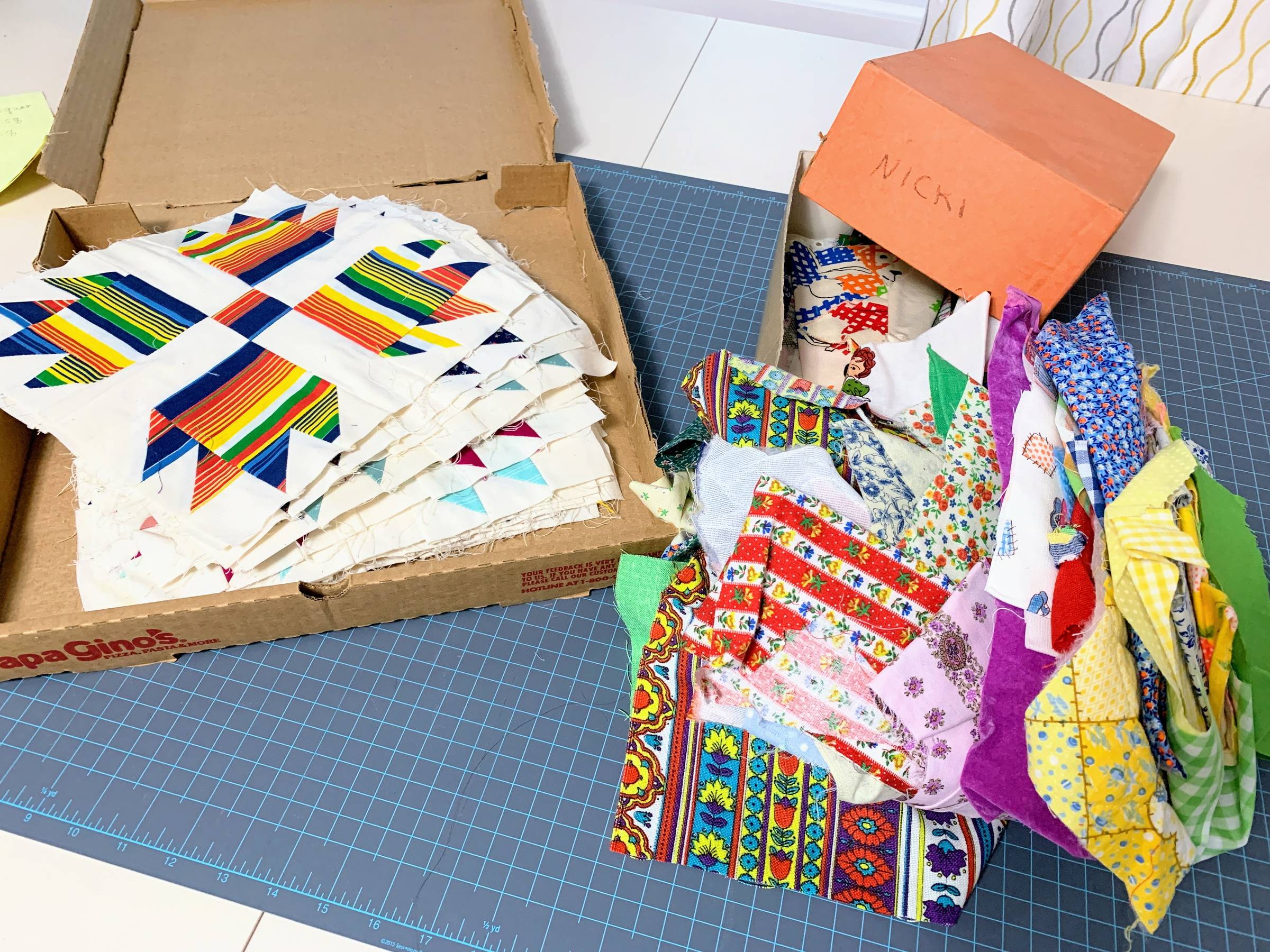 On the right, a pizza box with a stack of quilt blocks in it. On the left of the image, there is a box with and orange lid and scraps are flowing out of it.