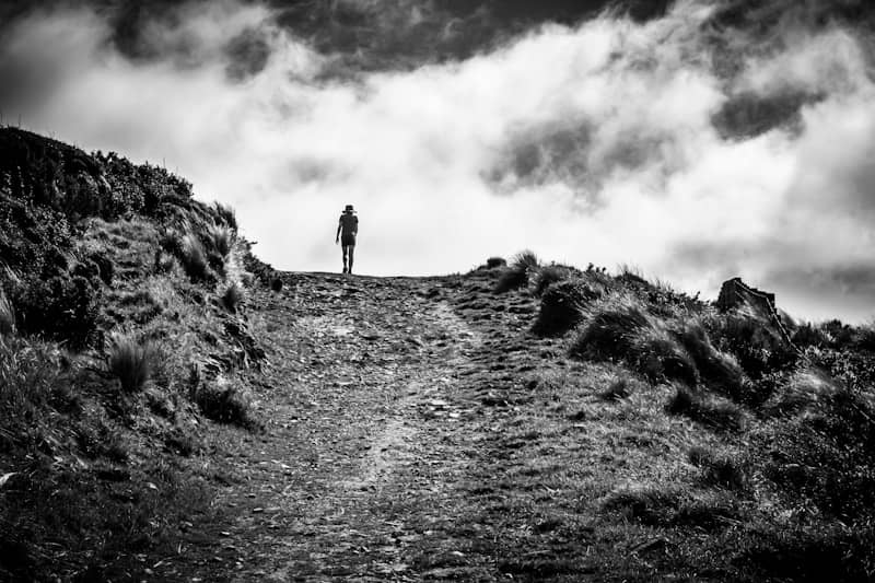 A lone figure walks up a grassy hill under clouds.