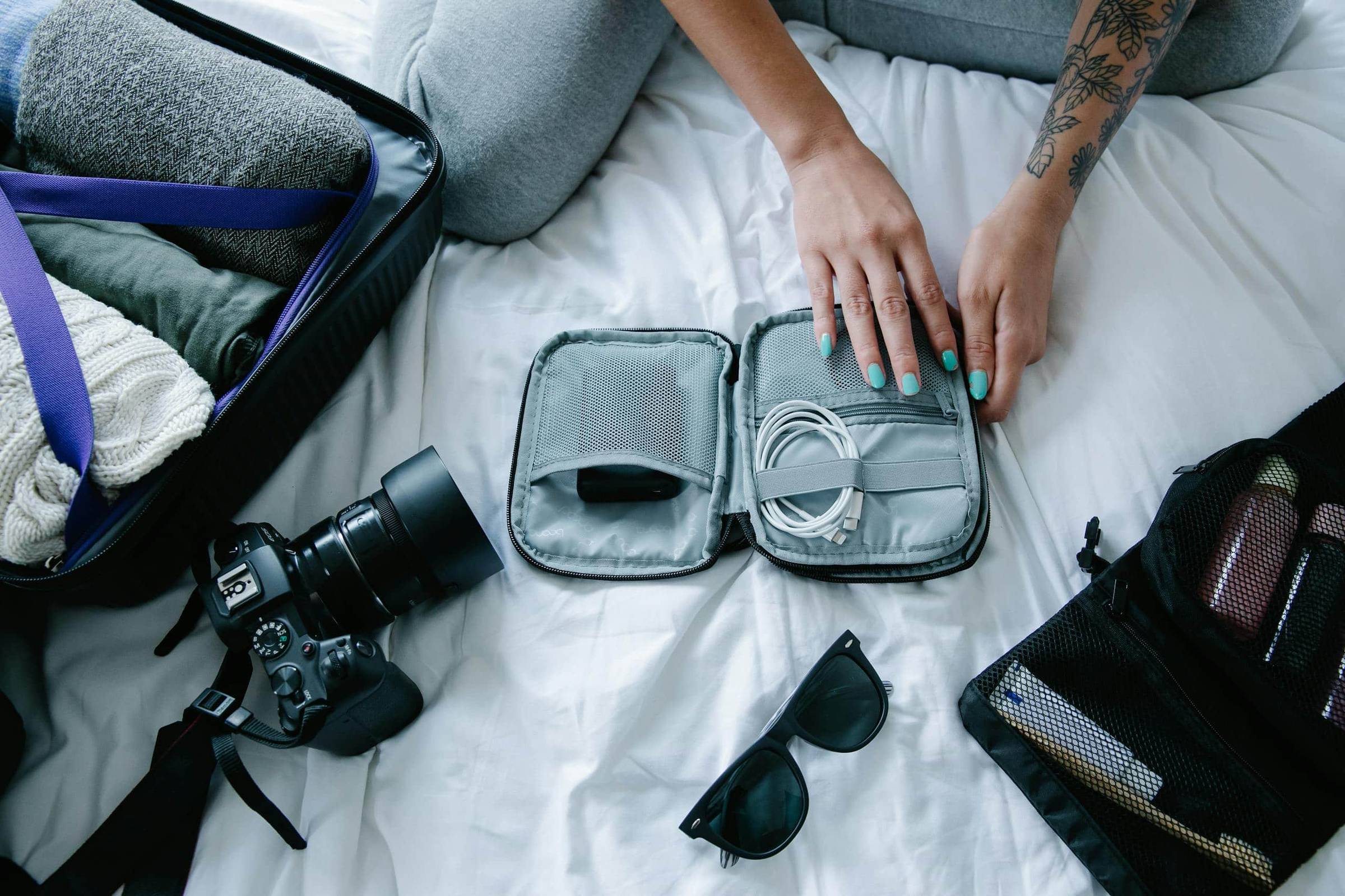 A person sits cross-legged on a bed while packing travel essentials, including an open suitcase with folded clothes, a small tech organizer holding charging cables and a power bank, a DSLR camera, sunglasses, and a mesh toiletry bag with travel bottles. Their hands, with teal nail polish and a floral tattoo on one arm, rest on the tech pouch.
