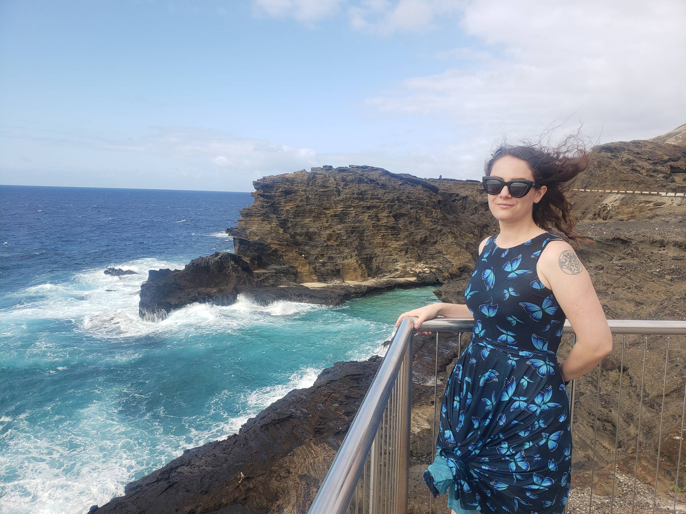 Person with long wavy hair wearing a blue butterfly-print dress and sunglasses stands at a seaside lookout on Oʻahu, Hawaiʻi. Behind them, turquoise waves crash against dark volcanic cliffs under a partly cloudy sky, capturing the beauty and power of the Pacific coastline.