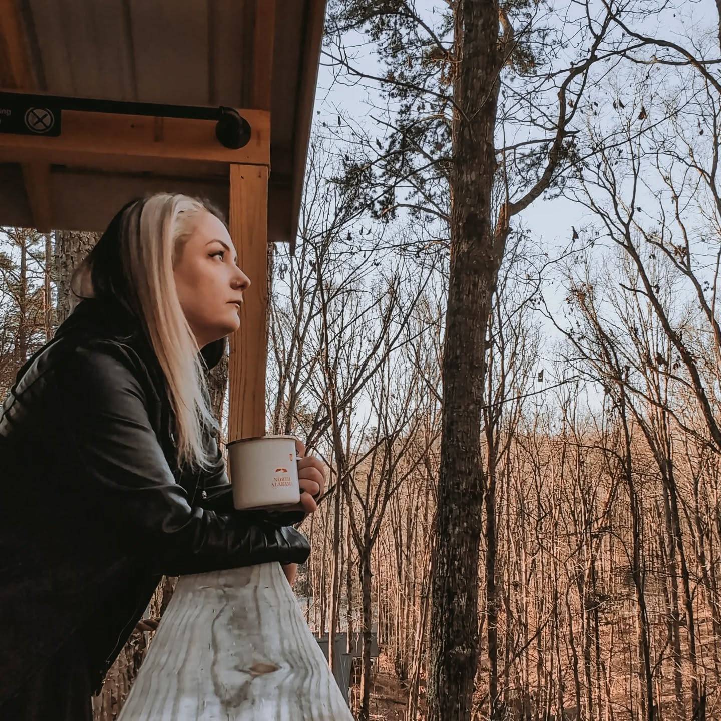 A woman enjoys a peaceful rest day on a cabin porch, leaning on the railing with a warm drink in hand, overlooking a quiet forest of bare trees.