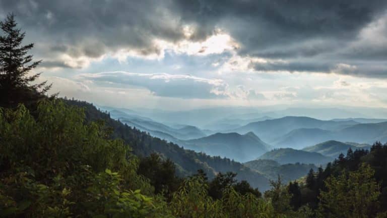 View of the Blue Ridge Mountains under a moody sky with sunbeams breaking through the clouds. Layers of misty blue ridges fade into the distance, creating a serene and peaceful atmosphere, perfect for those seeking quiet spaces.