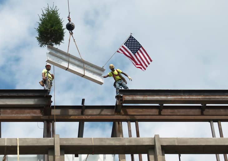 Image of a topping-out ceremony where a white steel beam with a tree and an American flag adorns it is being lowered into place. 