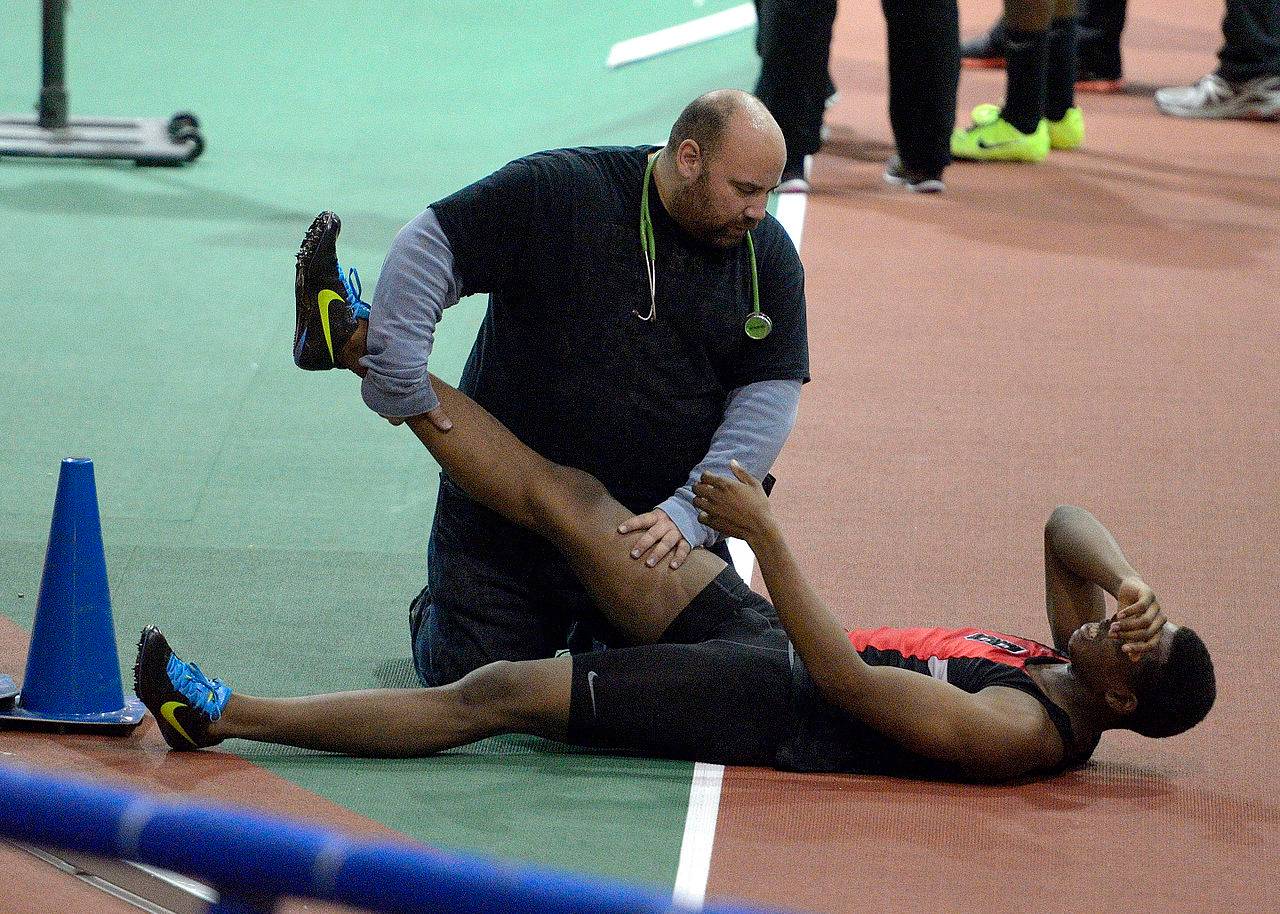 A track athlete lying on the ground with a medical staff holding his leg up in the air. His facial expression shows pain, suggesting his leg is cramping.