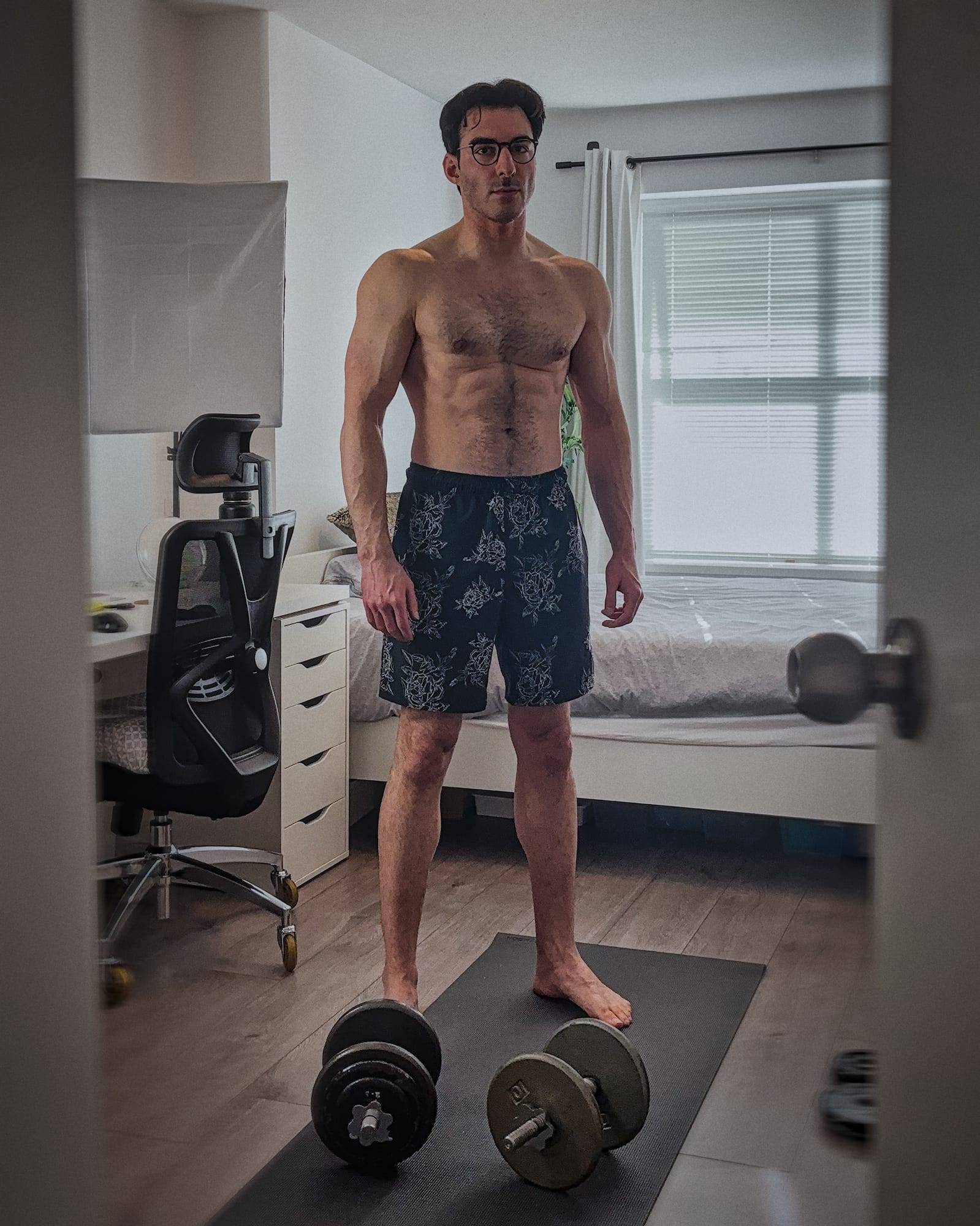 Poyan standing flexed in a bedroom with dumbbells on the floor.