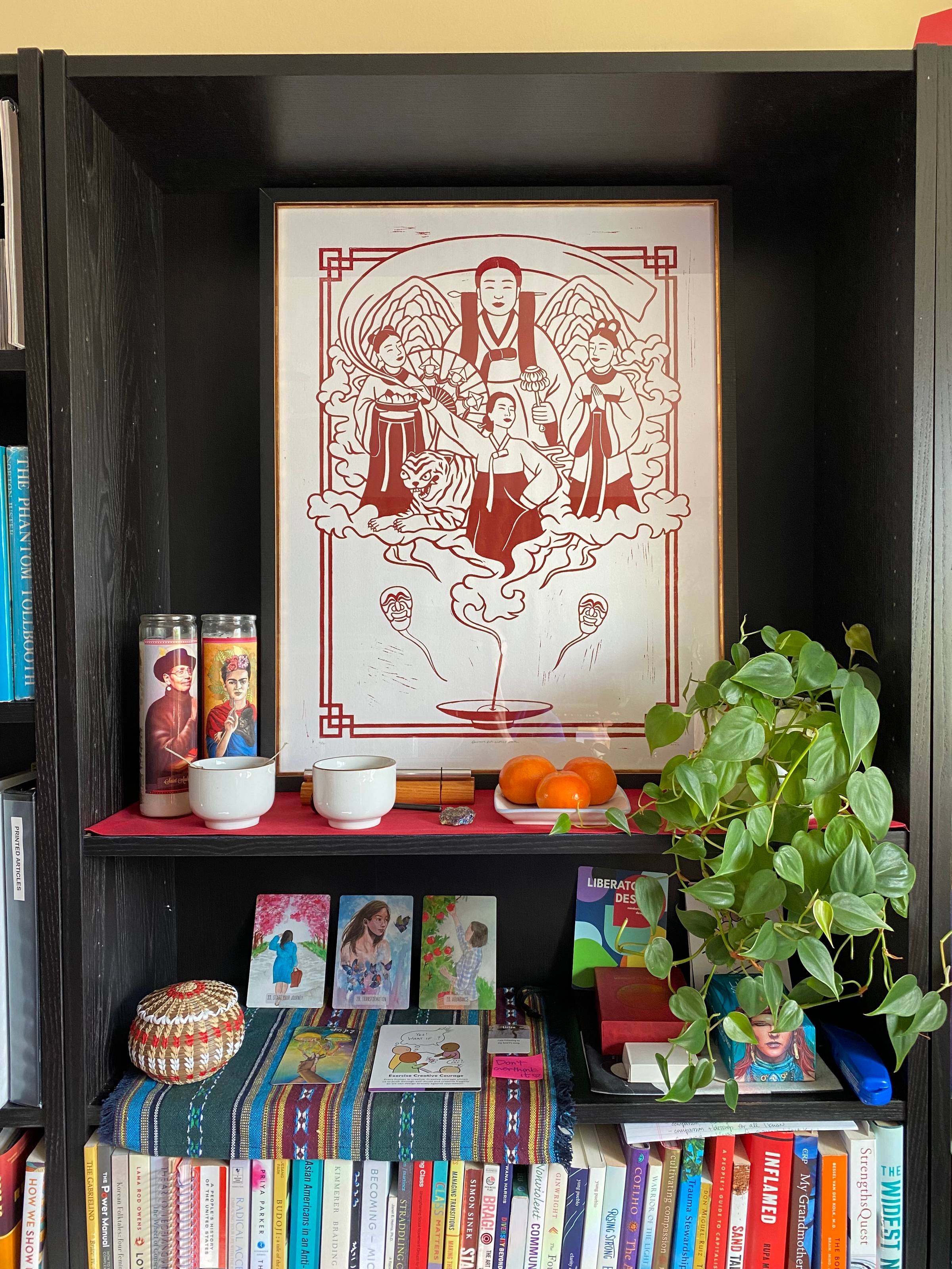 A home altar arranged on a black bookshelf. At the top center is a red and white woodblock-style print depicting Korean ancestral figures, including women in hanbok and a central figure holding ritual objects, surrounded by clouds and a tiger. Below are t