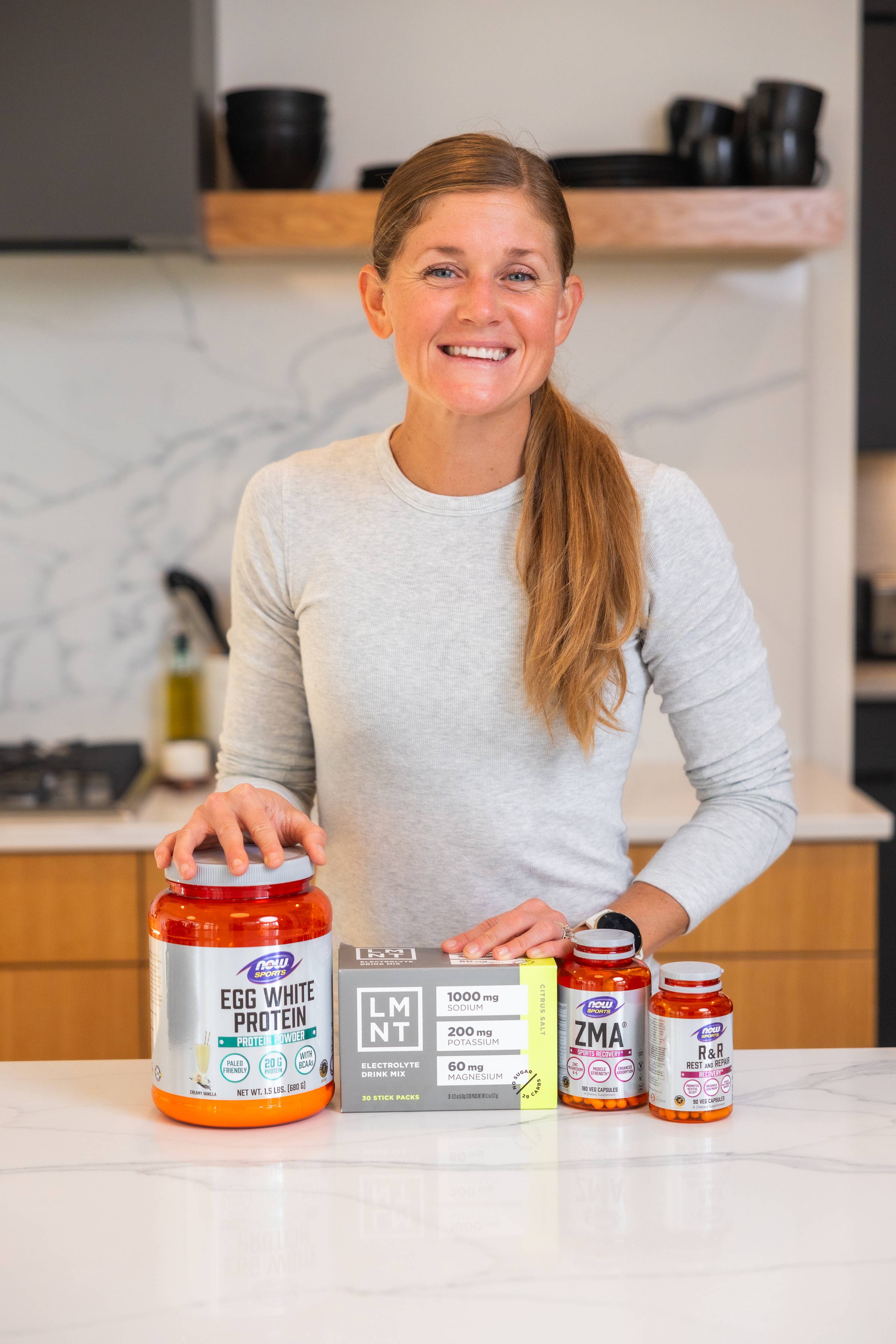 woman posing in her kitchen with protein powder, electrolytes and muscle recovery supplements