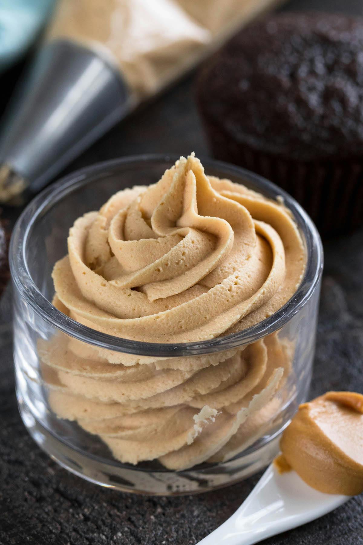 light brown frosting swirled into a short clear glass next to a spoonful of peanut butter.