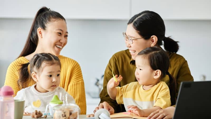 Two Asian moms sitting at a  table with their young daughters, laughing and talking
