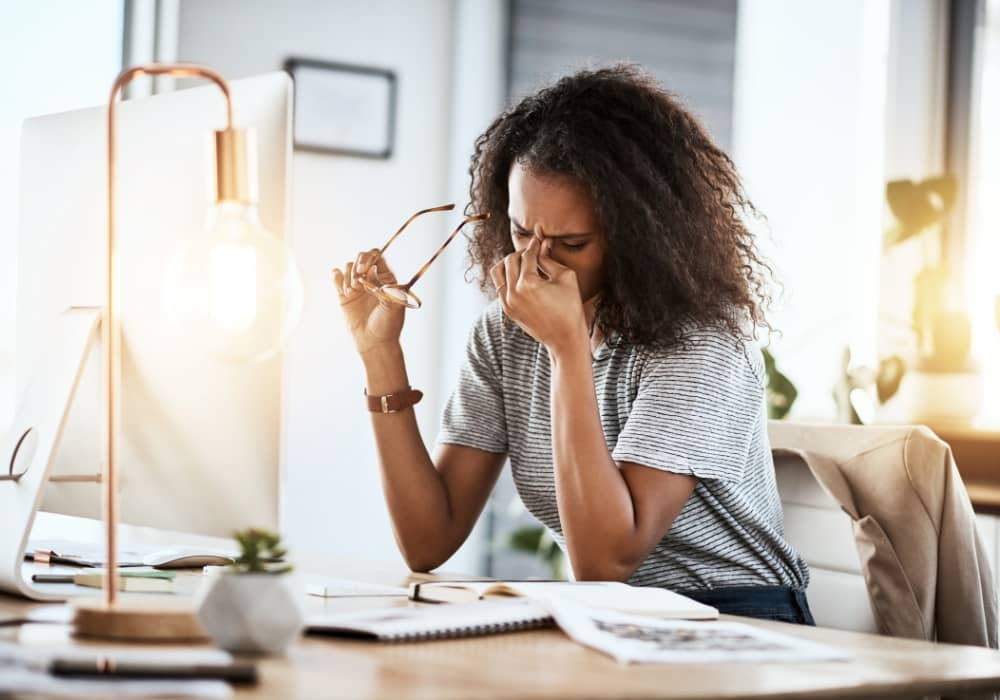 A Black woman in front of a computer, pinching her face and bowing her head