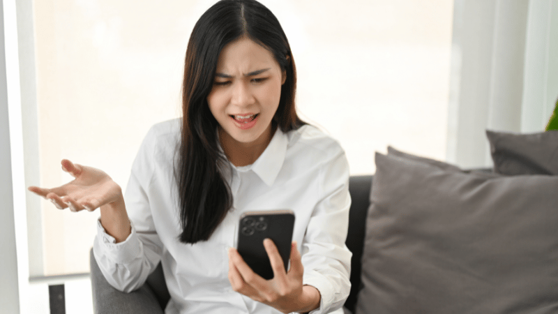 Photo of an “angry Asian woman” wearing a white shirt sitting on a dark couch, holding a cell phone in her left hand and talking angrily to it.