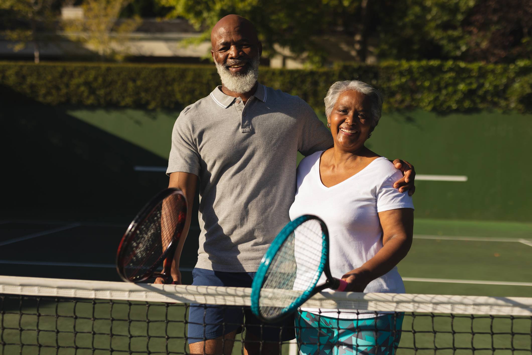 Older couple relaxing on a tennis court in front of the net holding rackets and smiling to the camera