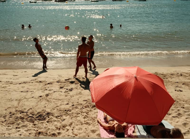 Sunny beach with people playing and a red sun umbrella.