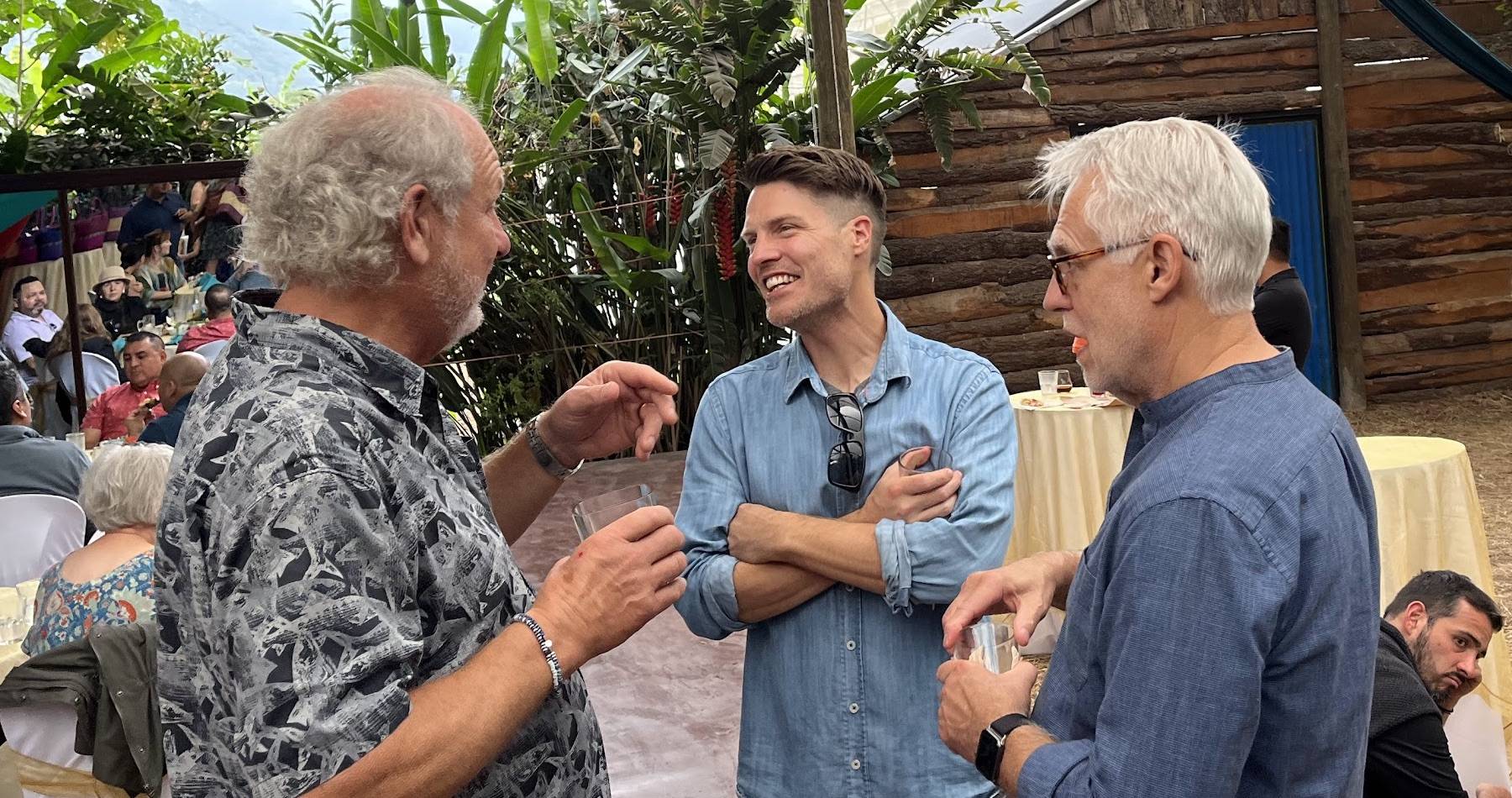 Photo: Justin Jackson (in the center, wearing a light blue shirt) smiling and engaged in conversation with two older men at an outdoor wedding reception. The setting appears to be a tropical venue with plants visible in the background. Justin is clearly energized and enjoying the social interaction.