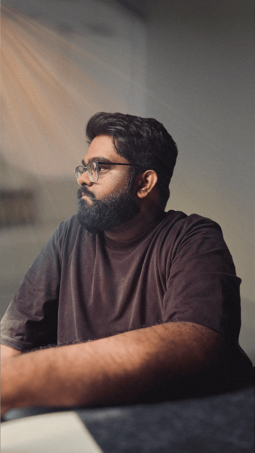 A man with a beard and glasses is sitting and looking to the side. He is wearing a dark brown shirt, and natural light is streaming in, creating soft rays visible in the background. The scene has a calm and introspective mood.