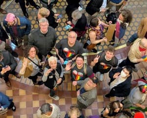 12+ white people many with rainbow heart shirts, looking up at camera above. Standing on mosaic floor of Iowa statehouse. 