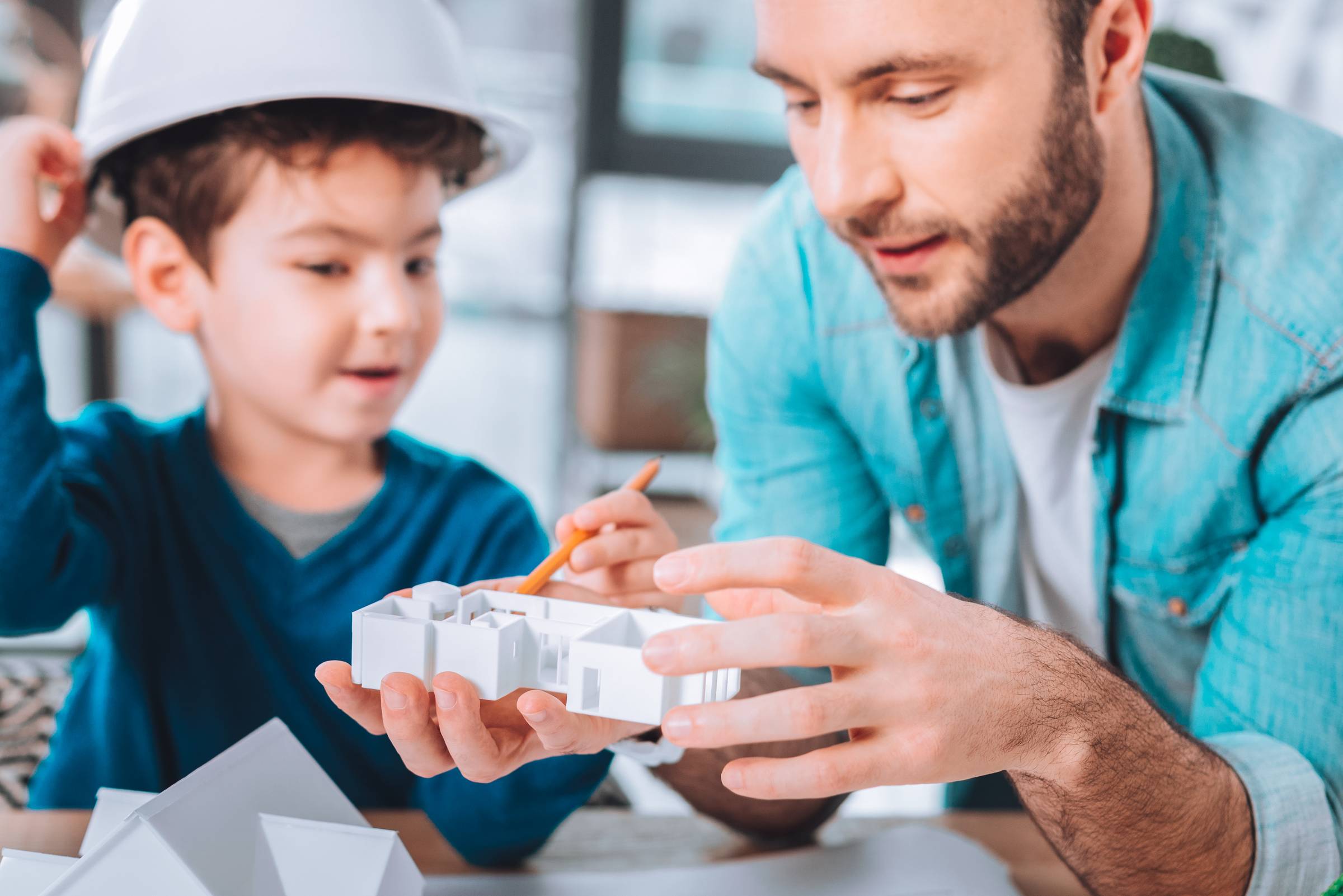 image of an adult male smiling while speaking about a handheld model home to a male child who is also smiling and wearing a white hardhat while pointing at the object with a pencil