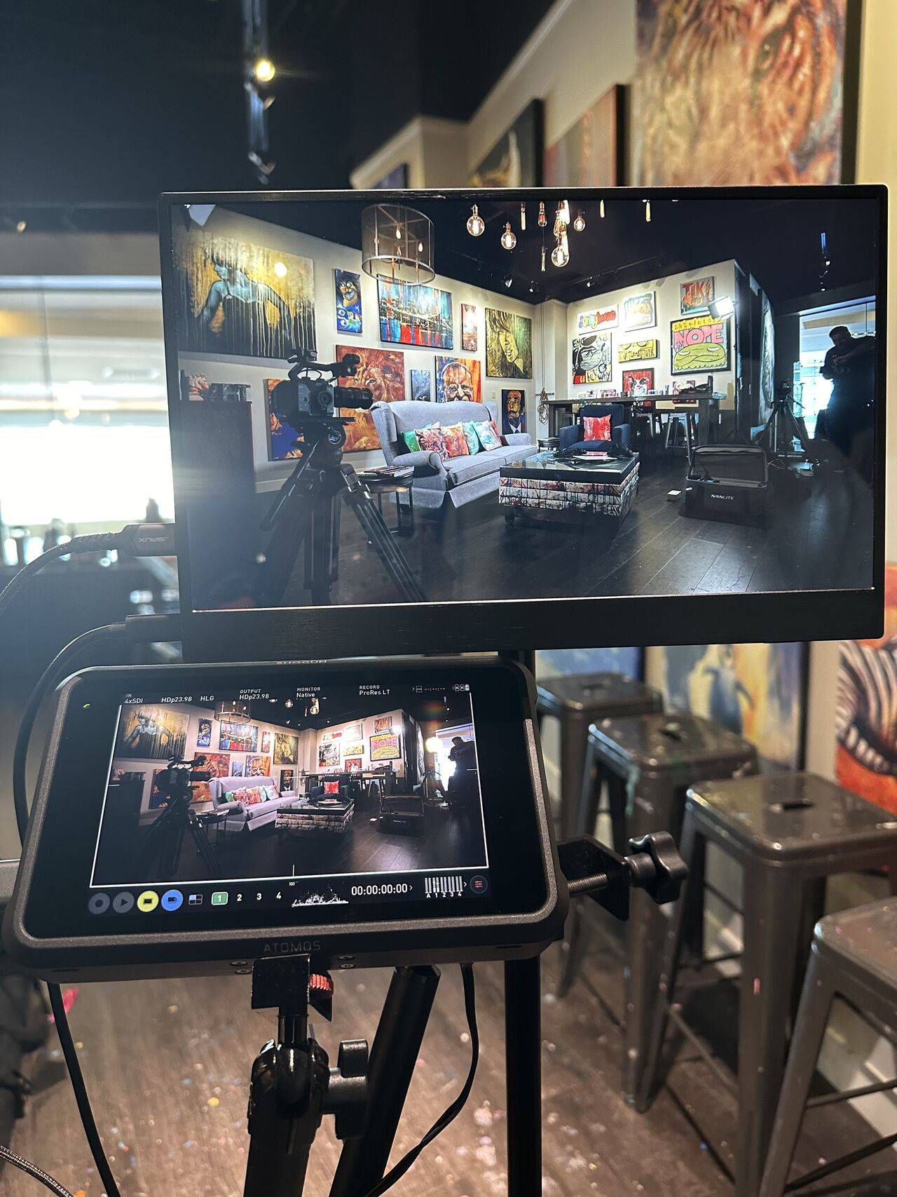 This image shows a behind-the-scenes look at a video shoot setup. It features two monitors on tripods capturing a cozy, art-filled studio space. The room being filmed has vibrant artwork displayed on the walls, a couch with colorful throw pillows, and other eclectic furniture. The lighting includes warm overhead bulbs, creating an inviting atmosphere. The monitor in the foreground displays the same scene, with a timer and recording settings visible on-screen.