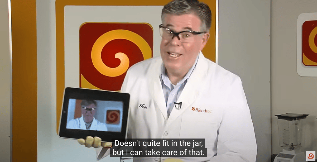 Man in a white lab coat labeled ‘Tom’ from Blendtec holds a tablet showing his own image. He smiles at the camera inside a demo room with Blendtec logos. Caption reads: ‘Doesn’t quite fit in the jar, but I can take care of that.’ 