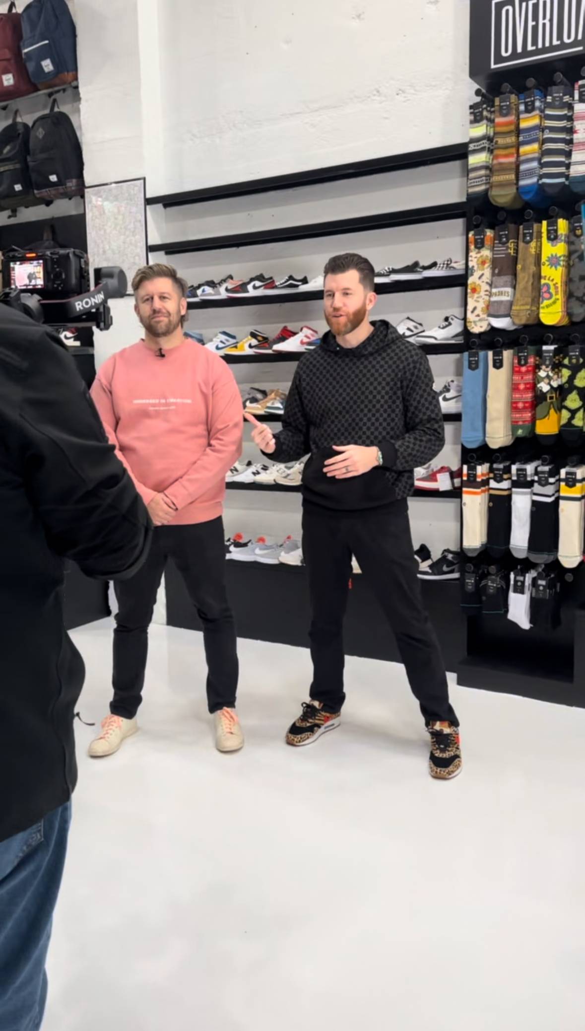 This image captures two men standing inside a sneaker and accessory store during what appears to be a filmed conversation or interview. One man is wearing a pink sweatshirt, while the other is dressed in a black hoodie and sneakers with animal print details. Behind them is a wall displaying various sneakers and socks, neatly organized. A cameraman with professional equipment is visible in the foreground, recording the scene.