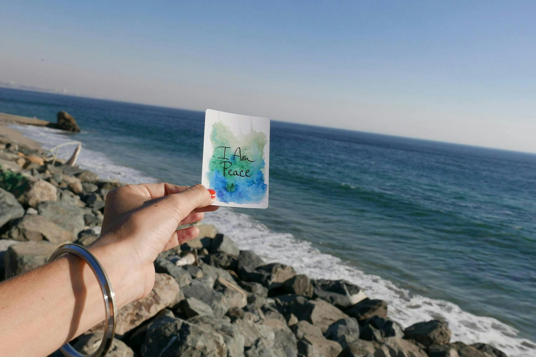 A woman standing in front of water at a beach, holding a piece of paper with the affirmation "I am peace" written on it