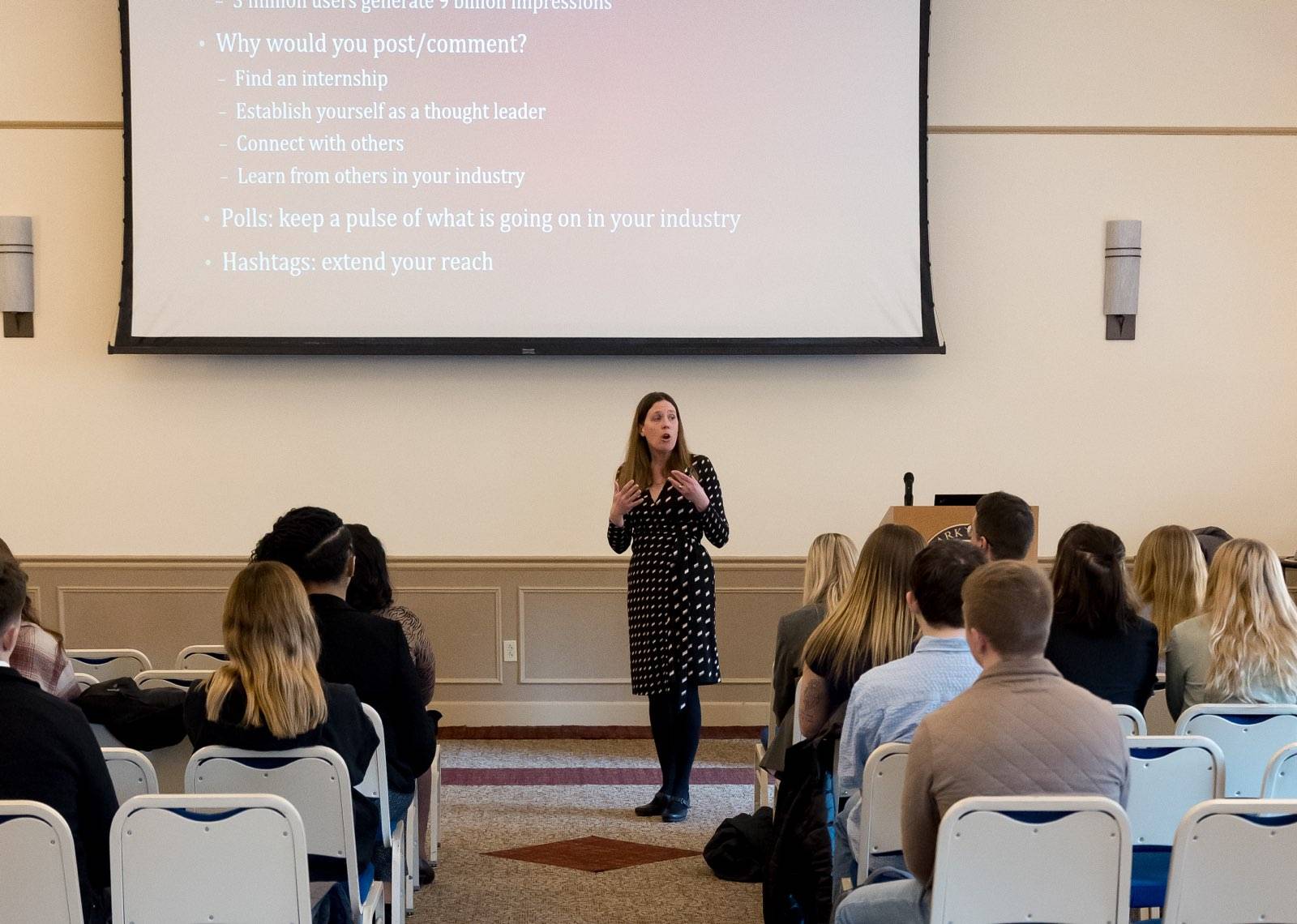 A woman stands at the front of a classroom giving a presentation to a group of seated attendees. She is wearing a black dress with white patterns. Behind her, a large screen displays text about the benefits of posting and commenting online for professiona