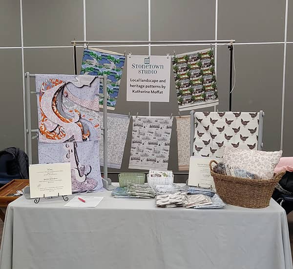 A table at a craft show displays tea towels and throw pillow covers from Stonetown Studio. 