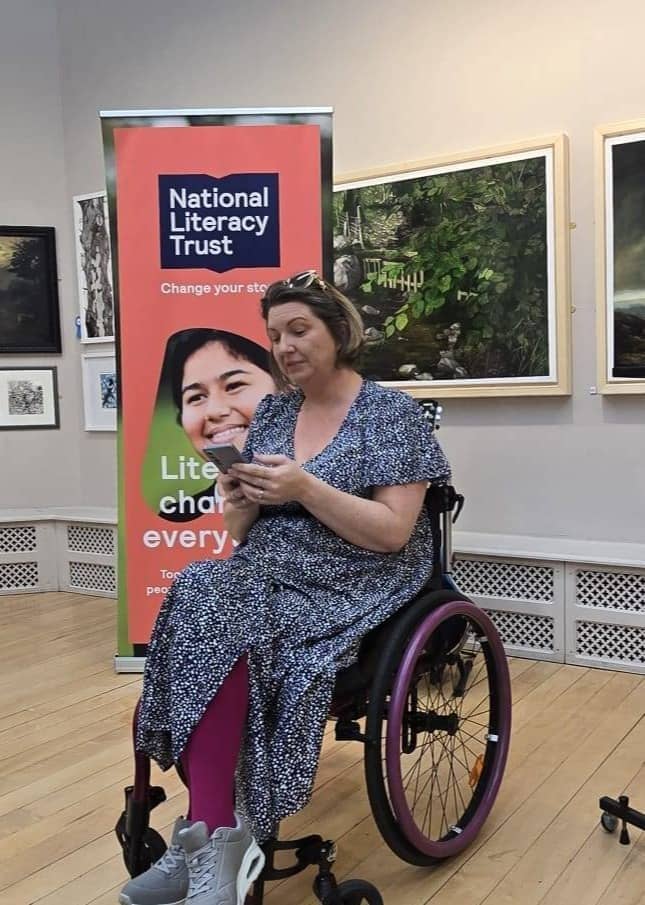 Woman in wheelchair reading from her phone. Wearing a blue dress with fine white spot pattern. The banner in the background reads The National Literacy Trust
