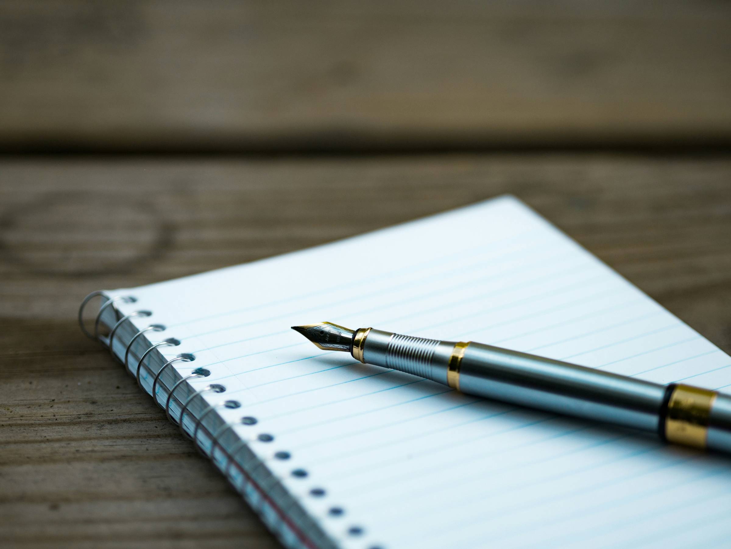 A lined notepad with a fountain pen on top. The notebook sits on a bench type tabletop.