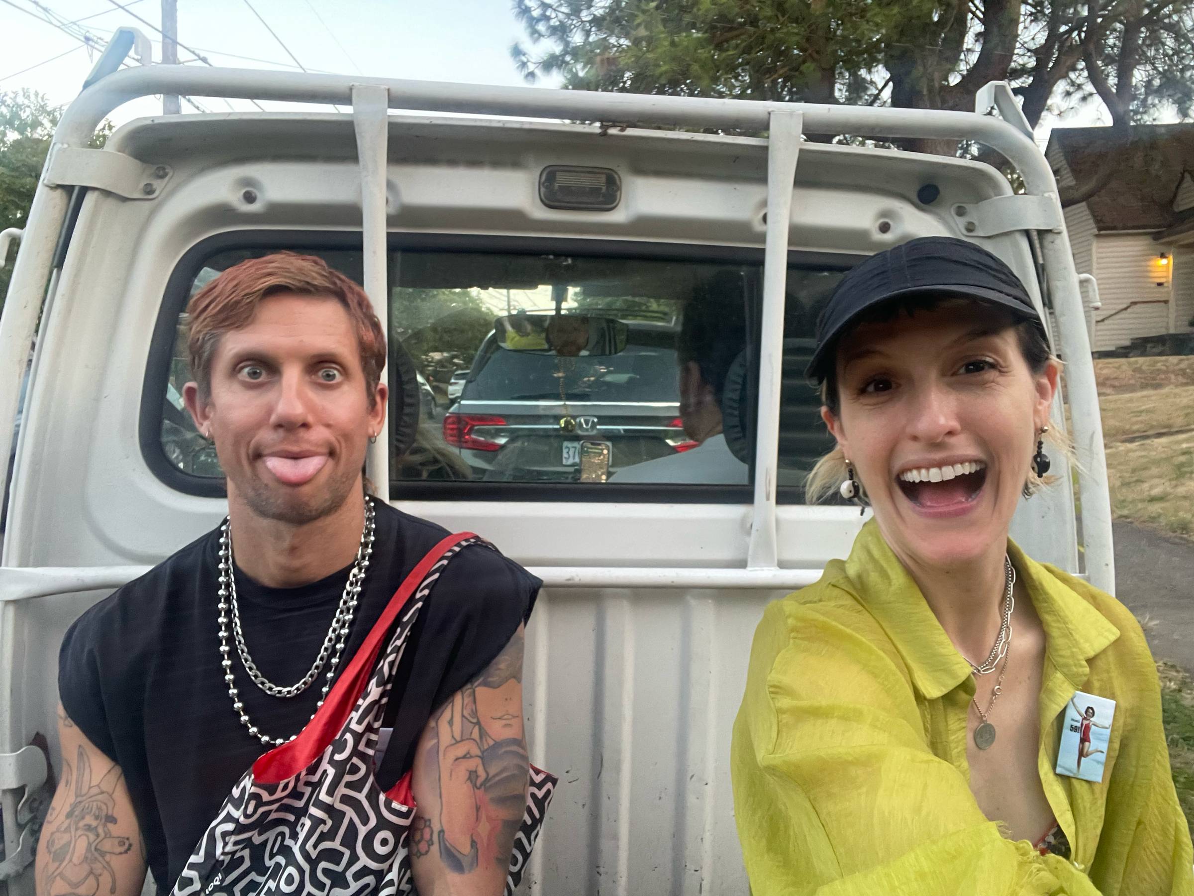 A man and a woman pose for a selfie in front of the tailgate of a white pickup truck, parked on a residential street with trees and a house visible in the background. The man, on the left, has reddish-brown hair and tattooed arms, sticks out his tongue, and wears a black t-shirt with layered silver chain necklaces and a red-and-black patterned crossbody bag. The woman, on the right, wears a black cap and an open yellow-green shirt over a red top, with layered necklaces and dangly earrings, and laughs with her mouth wide open. A dark SUV with its rear window visible is parked behind them.