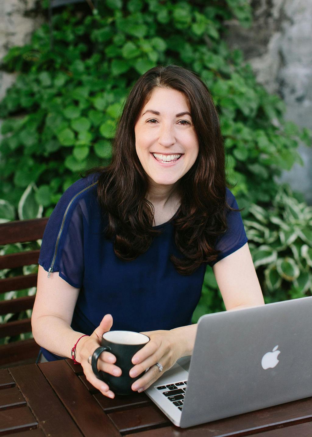 Bev sitting at a table outside with her laptop in front of her. She is smiling and has a mug in her hand. She is wearing a dark blue shirt and has long dark brown hair. There is a green tree behind her.