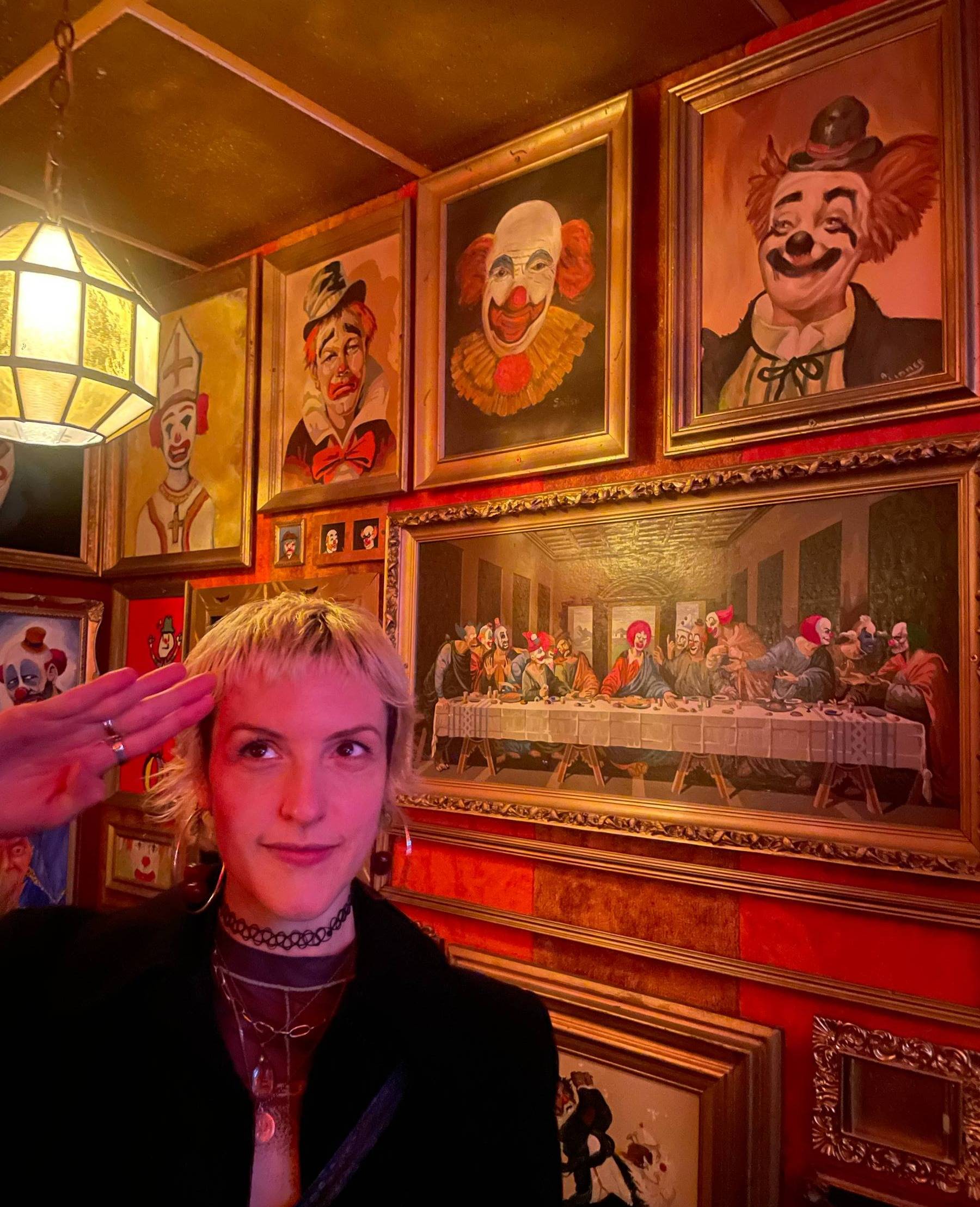 A picture of Devin with short blonde hair, a choker necklace, and a black jacket, saluting the camera, in a dimly lit red-and-gold room covered with paintings of clowns, including a clown-themed parody of "The Last Supper."