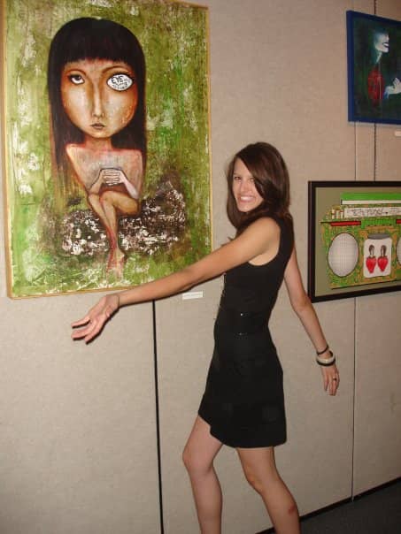 Young Devin in a black dress wearing a huge smile. She is gesturing to a large painting on a gallery wall. The painting is of a girl with her hands folded in front of her on a green background. 