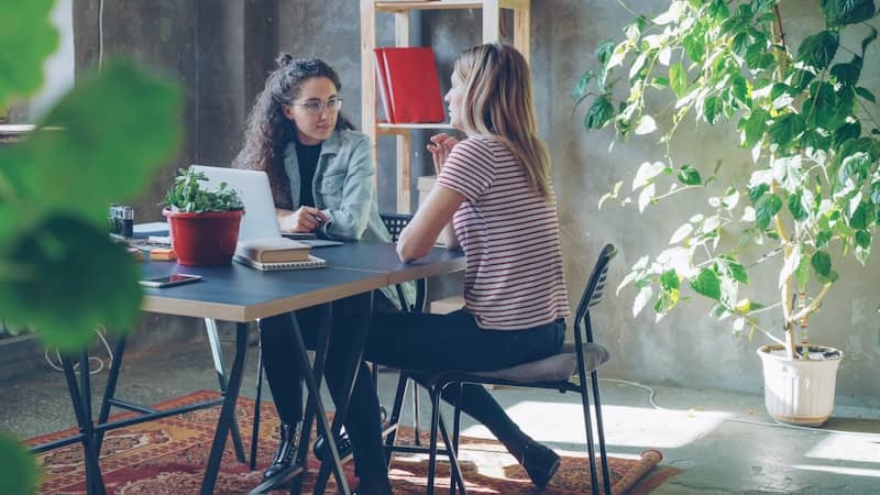 Two women are sitting at a desk, conversing.