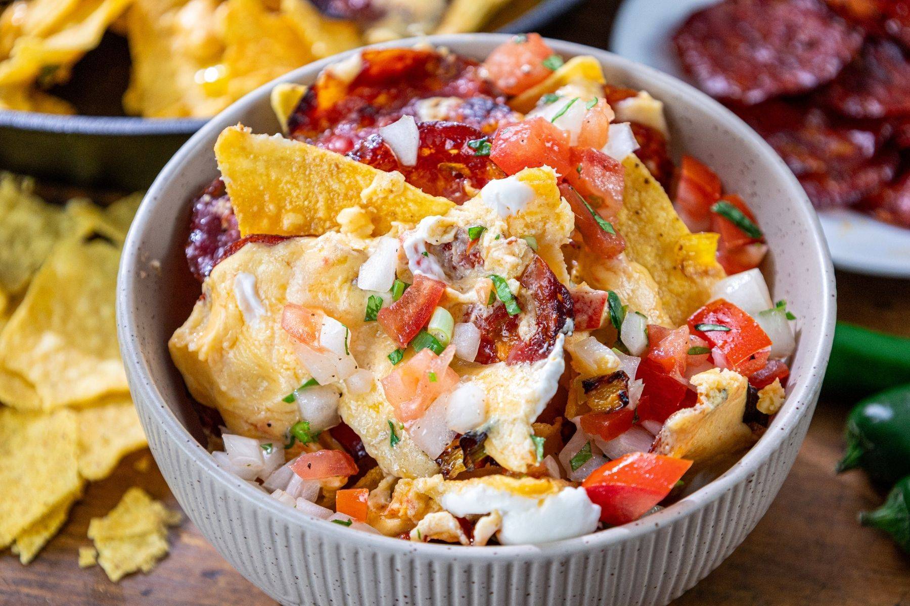 A bowl of nachos topped with melted cheese, sliced chorizo, diced tomatoes, onions, and cilantro, with tortilla chips visible around the bowl and in the background.