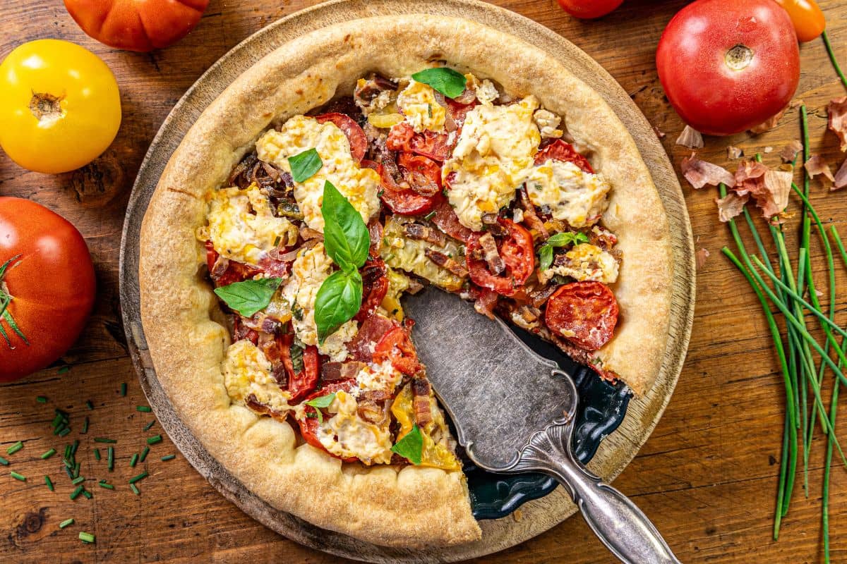 A deep-dish pizza topped with sliced tomatoes, pepperoni, cheese, and fresh basil sits on a wooden plate. A metal pie server holds one slice, and fresh tomatoes and chives are scattered on the wooden table.