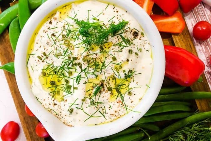 A bowl of creamy white dip garnished with fresh dill, chives, black pepper, and olive oil, surrounded by assorted fresh vegetables including green beans, red bell pepper, cherry tomatoes, and snap peas.