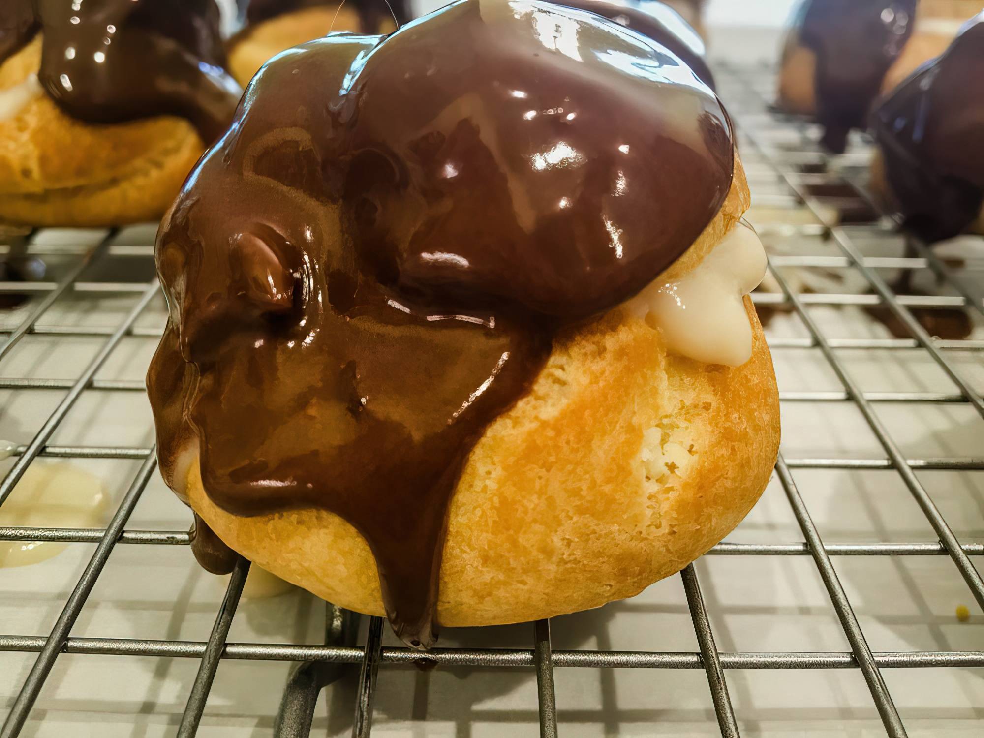 A close-up of a cream puff on a cooling rack, filled with vanilla cream and topped with glossy chocolate glaze. Cream is oozing slightly from the side. Other pastries are blurred in the background.